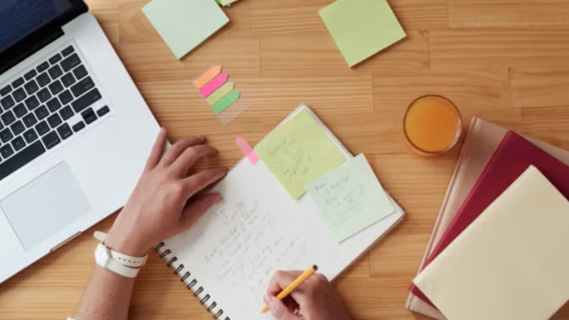 Overhead view of hands writing in a notebook surrounded by sticky notes and a laptop.