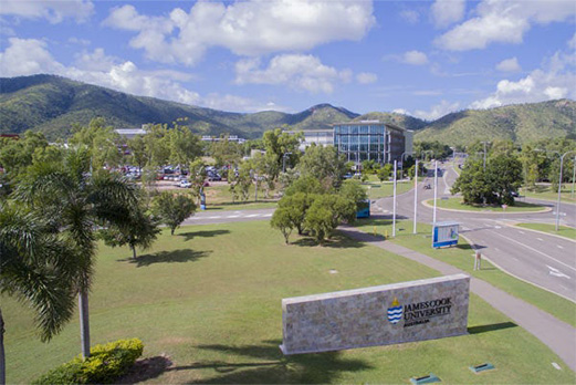Aerial view of James Cook University’s Townsville campus featuring modern buildings, tree-lined roads, parked cars, and scenic mountain ranges in the background under a partly cloudy sky.