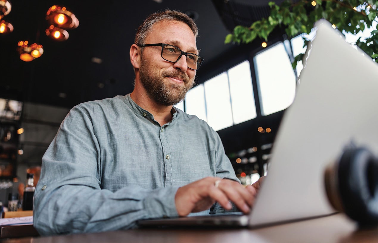 A bearded man in glasses using a laptop in a café, smiling, with a gray background.
