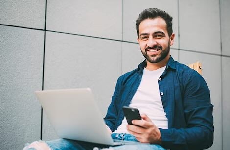 Student sitting with laptop and phone, smiling at camera.