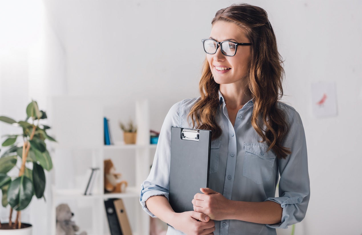 A smiling woman wearing glasses holding a clipboard in a bright, indoor office space.
