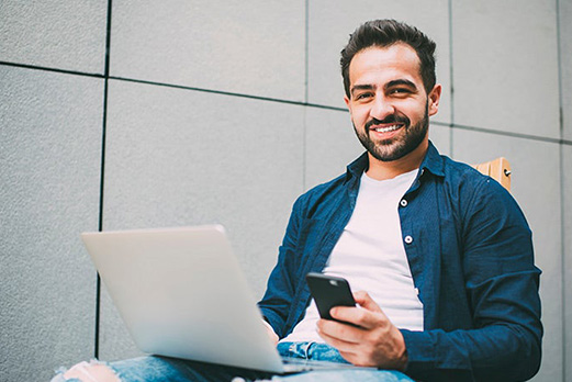 Man smiling while working on a laptop and holding a smartphone, seated outdoors against a modern tiled wall.